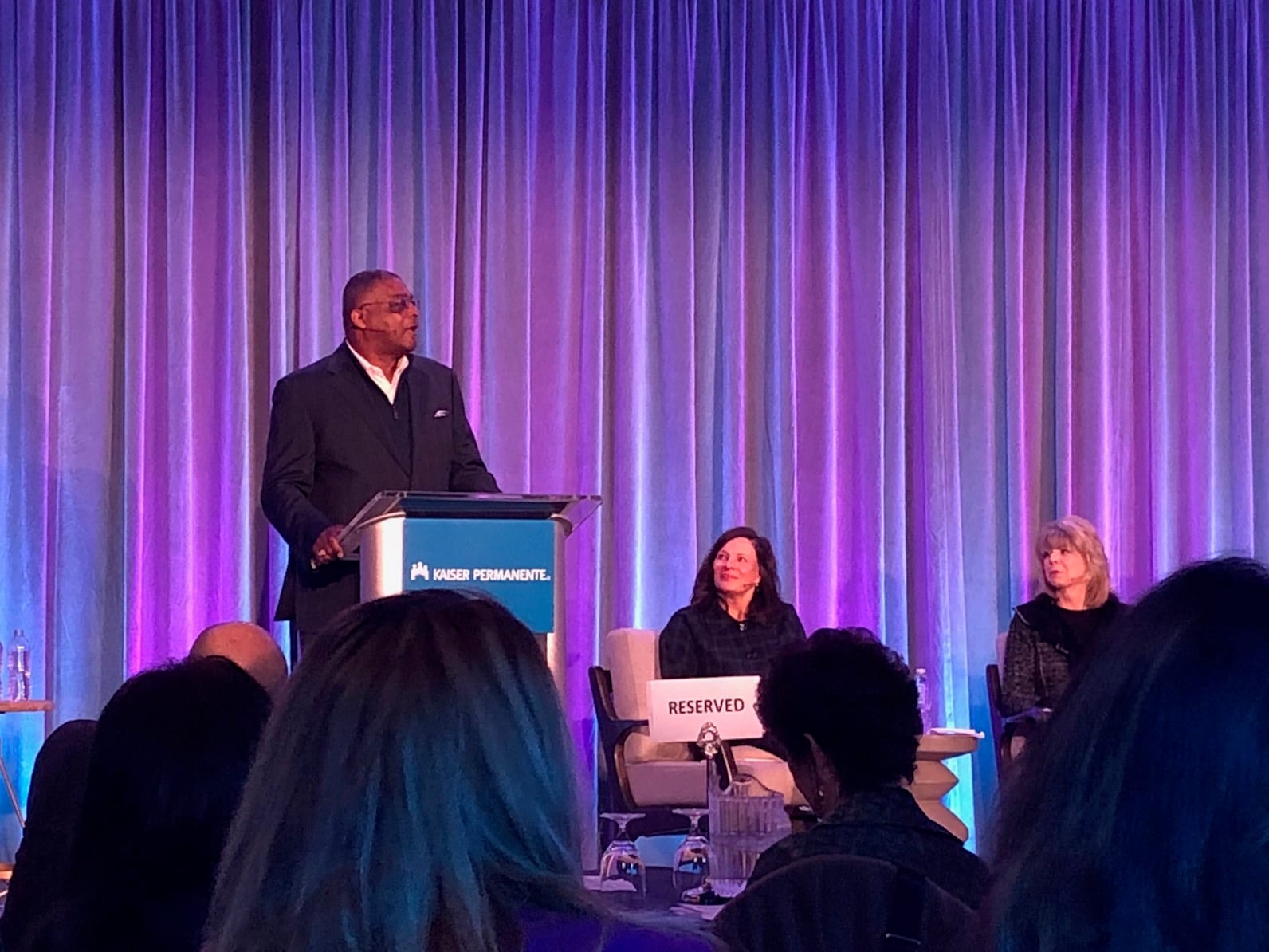 Dennis Dabney at the podium beside a panel on a stage with purple curtains